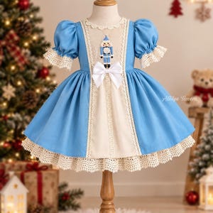 May include: A light blue and cream dress featuring puff sleeves, a central cream panel with a nutcracker embroidery, a white bow at the waist, and intricate cream lace trim along the neckline, sleeves, and hemline. The text Atolye Tanem is visible.