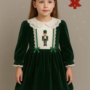 Op de afbeelding: Een groene fluwelen jurk met een witte kanten kraag en boord. De jurk heeft lange mouwen en een notenkrakerontwerp op het lijfje, met groene strikken. Een rode kerstboomversiering hangt op de achtergrond.