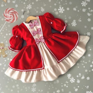 以下が含まれることがあります： 白と赤のクリスマスドレス。白いレースのトリムと赤と白のストライプのリボンが付いています。ドレスはパフスリーブで、スカートはボリュームがあります。