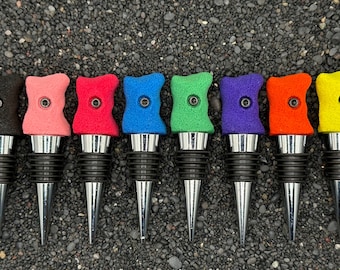 May include: A collection of seven colorful wine bottle stoppers. Each stopper features a unique, textured top in black, pink, red, blue, green, purple, orange, and yellow. The stoppers have a silver base and a black rubber seal.