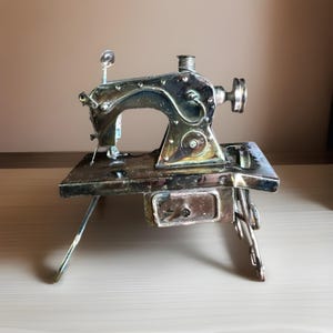 May include: A vintage silver-toned sewing machine figurine with intricate details. The miniature machine features a needle, spool holder, and a small drawer. It is set on a table with three legs, showcasing a classic design.