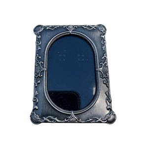 May include: A rectangular silver-toned metal picture frame with an oval-shaped center opening. The frame features intricate embossed scrollwork and shell-like motifs at each corner and along the edges, surrounding a dark, reflective glass pane.