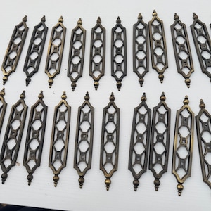 May include: A collection of vintage bronze-toned decorative hardware pieces. Each piece is a long, rectangular shape with an openwork geometric design and a decorative finial at the top and bottom. The hardware is arranged on a white surface.