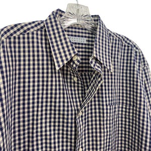 May include: A long-sleeve button-down shirt featuring a navy blue and white gingham check pattern. The shirt includes a pointed collar, a front button placket with white buttons, and a chest pocket. A label reads Barneys New York at the neck.