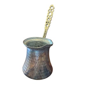May include: A hammered metal coffee pot with a wide base, a narrow neck, and a flared rim. The pot features a long, upright handle with an intricate, openwork geometric pattern. The exterior surface displays a weathered, dark, and mottled patina.