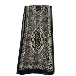 May include: A rectangular scarf featuring a sheer black base with an intricate gold-toned filigree pattern. The design includes a central medallion motif surrounded by ornate scrolling borders. The brand name Elaine Gold is printed in the corner.