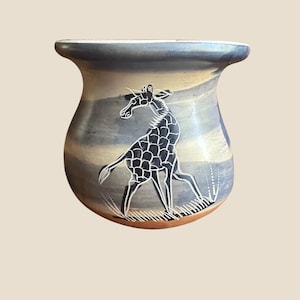 May include: A rounded ceramic pot featuring a wide, flared rim and a smooth, multi-toned surface in shades of blue, gray, and cream. The exterior is decorated with a detailed, white-etched graphic of a giraffe standing on stylized grass.