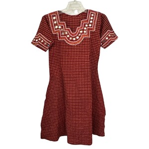 May include: A short-sleeved dress in a dark red and white grid pattern. The neckline and sleeve cuffs feature a decorative zigzag border with embedded circular mirrors and white stitching, creating a distinct contrast against the red fabric.