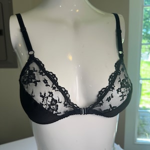 May include: A black bra featuring sheer lace cups with a floral pattern and a solid black lower band. The garment includes thin, adjustable black shoulder straps and a small center-front closure mechanism connecting the two cups together.