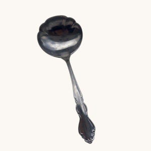 May include: A silver-colored ladle with a scalloped bowl and a long, ornate handle. The handle features decorative detailing near the base and a smooth, curved design. The ladle is designed for serving soups and sauces.