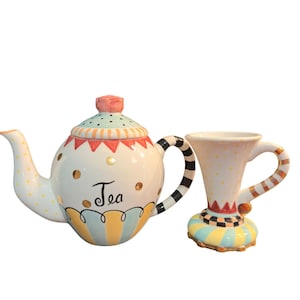 May include: A ceramic teapot and matching mug featuring a cream-colored base with gold polka dots, a light blue and yellow striped bottom, and black and white striped handles. The teapot lid has a pink rose finial and the word Tea is written in black.