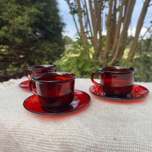 May include: Three translucent deep red glass teacups with matching saucers. Each cup features a rounded bowl shape and a small side handle. The set is arranged on a textured off-white surface, showcasing the vibrant ruby-colored glass material.