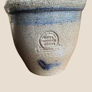 May include: A tan stoneware crock with a speckled texture, featuring a blue band near the rim and a blue painted leaf motif near the base. The front displays a circular stamp reading HAND CRAFTED ROWE POTTERY WORKS CAMBRIDGE WI 1995.