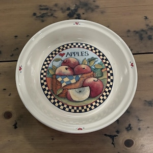 May include: White ceramic bowl with a decorative apple design. The bowl features a central image of a basket filled with red apples, a blue and white checkered cloth, and the word "APPLES" above. The rim has small heart accents.