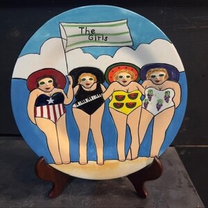 May include: A decorative ceramic plate featuring four women in swimsuits, each with a different design. The plate has a blue sky and ocean background, with a banner that reads "The Girls". The plate is displayed on a wooden stand.