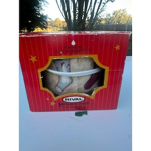 May include: A red rectangular Rival Potpourri Crock box with vertical white stripes and yellow stars. The front features a cutout window displaying a ceramic crock decorated with a snowman on a blue stocking and text reading Perfect for liquid or gel potpourri.