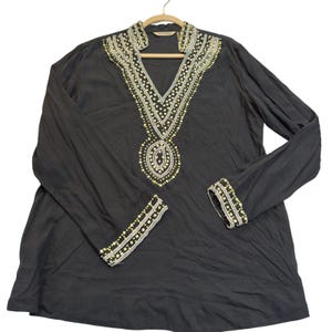 May include: A black long-sleeved tunic featuring a deep V-neckline adorned with intricate silver and gold beadwork. The same metallic beaded pattern is repeated on the cuffs of the sleeves, creating a cohesive design on the dark fabric.