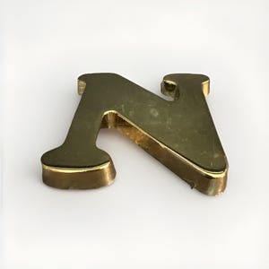 May include: A three-dimensional letter N crafted from a reflective gold-toned metal. The letter features a smooth, polished surface with a thick profile and distinct serif-style ends on the vertical strokes, creating a bold, metallic appearance.