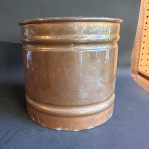 May include: A cylindrical copper bucket with a polished metallic finish and a warm, reddish-brown hue. The vessel features two horizontal raised bands encircling the body, providing structural detail to the smooth, reflective surface of the metal.