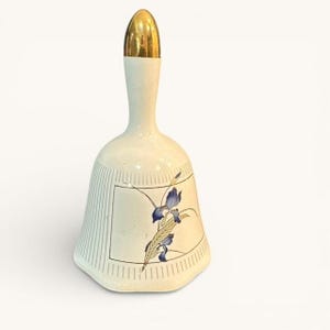 May include: Cream-colored ceramic bell with a gold-colored top. The bell features a rectangular design with vertical lines and a floral illustration in blue and gold. A decorative item.