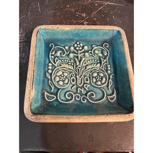 May include: Square ceramic dish with a turquoise interior and a raised floral design in white. The dish has a light brown, textured border. The floral design includes stylized flowers, leaves, and swirls.