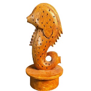 May include: A light brown wooden seahorse sculpture featuring a series of small, evenly spaced circular perforations across its body. The seahorse is mounted on a solid, cylindrical wooden base, showcasing a natural wood grain texture throughout.