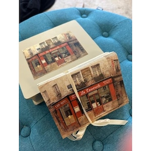 May include: A set of notecards and a matching box featuring a vintage illustration of a French cafe, "La Casserole." The cafe has a red awning and door. The cards are tied with a cream-colored ribbon, and the box is beige.