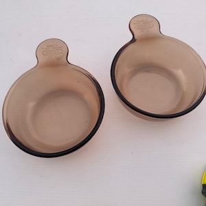 May include: Two Vision brand glass bowls with a brown tint and black rims. Each bowl has a handle and a rounded shape. The bowls are empty and appear to be in good condition. The bowls are likely for cooking or serving.