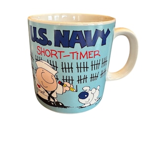 May include: A light blue ceramic mug featuring the text U.S. NAVY SHORT-TIMER in dark blue and red. The design includes a cartoon sailor marking tally marks on a wall with a pencil, accompanied by a small white dog with a blue nose.