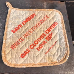 May include: A square, quilted off-white potholder with a yellow-orange fabric border and a hanging loop. The text in orange reads: SAVE ENERGY, WHITE-WESTINGHOUSE, GAS CLOTHES DRYERS have ESP, and Energy Savings Performance.