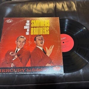 May include: A white ceramic mug with a black handle and the text "But First, Coffee" in a bold, black font. The mug is set against a blurred background, suggesting a cozy setting.