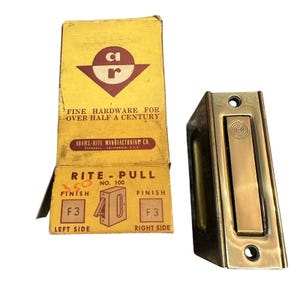 May include: A brass flush door pull hardware piece with a rectangular face and a central retractable handle, accompanied by its original yellow cardboard box labeled Adams-Rite Manufacturing Co. Rite-Pull No. 100 with F3 finish details.