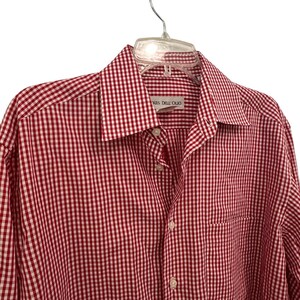 May include: A long-sleeved button-down shirt featuring a red and white gingham check pattern. The shirt has a pointed collar, a front button placket, and a chest pocket. A label inside the collar displays the text Louis Dell'Olio.