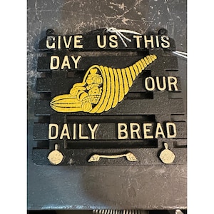 May include: Black metal wall plaque with the words "GIVE US THIS DAY OUR DAILY BREAD" in white lettering. A yellow cornucopia filled with produce is in the center. Includes a small handle and two decorative elements.