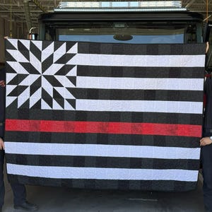 Könnte beinhalten: Ein rechteckiger Quilt mit einem schwarz-weißen Sternmuster in der oberen linken Ecke und horizontalen schwarz-weißen Streifen auf dem restlichen Stoff. Ein einzelner horizontaler roter Streifen verläuft durch die Mitte des Quilts.