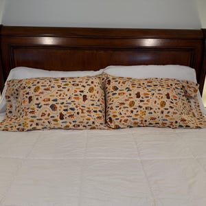 May include: Two decorative pillowcases with a beige background and a repeating pattern of bears, trees, and other outdoor elements. The pillowcases are on a white bed with a dark brown headboard.