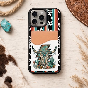 May include: Black phone case with a southwestern design. The case features a bull skull with turquoise accents, cacti, and flowers. The background has a cowhide pattern and a leather card holder.