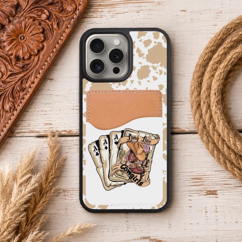 May include: A black phone case with a brown leather card holder and a white and tan cowhide pattern. The case features a graphic of playing cards, a cowboy hat, and the text "Long Live Cowgirls."