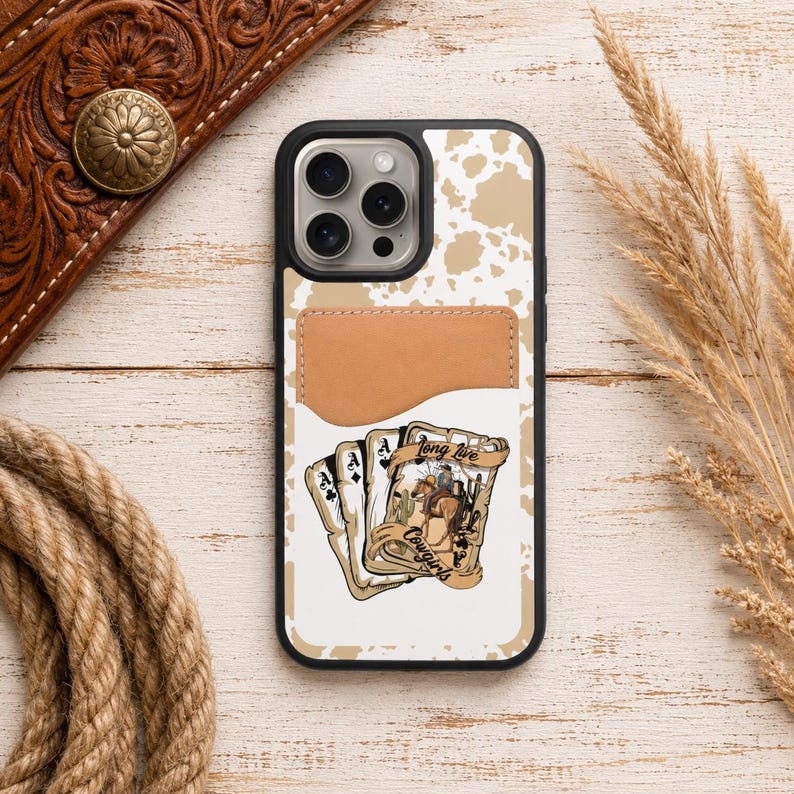 May include: A black phone case with a white and tan cowhide pattern and a brown leather card holder. The case features a graphic of playing cards with a cowboy theme and the text "Long Live Cowgirls".