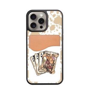 May include: A black phone case with a tan cowhide pattern and a tan leather cardholder. The case features a graphic of playing cards with a cowboy hat and the text "Long Live Cowgirls."