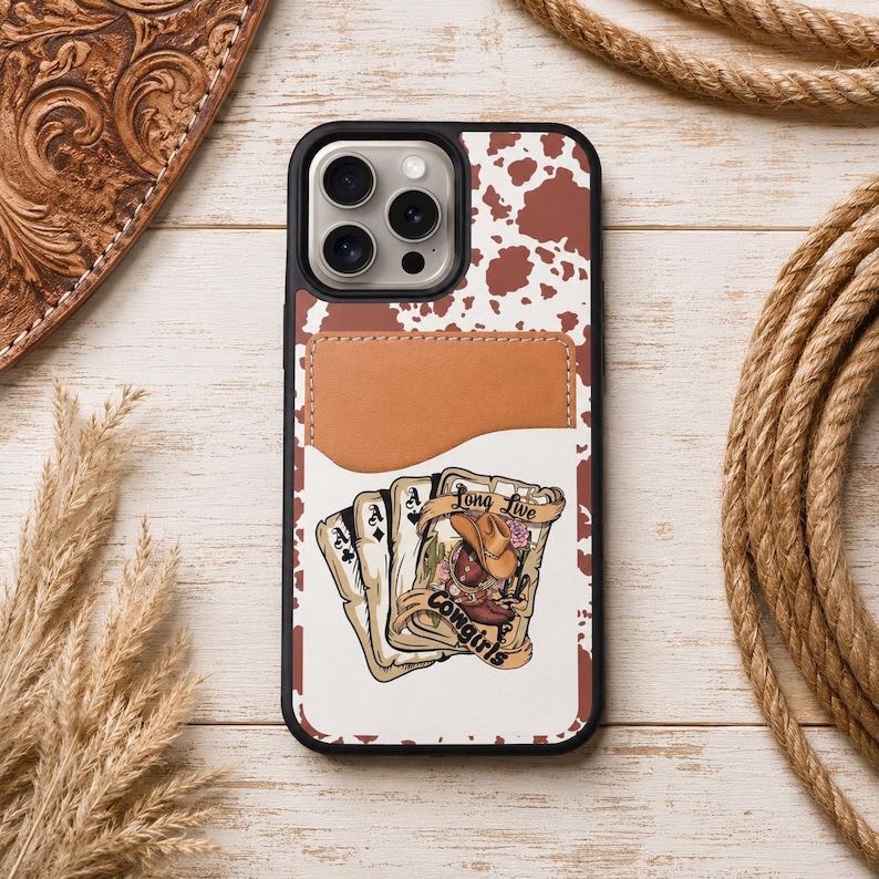 May include: A black phone case with a brown and white cowhide pattern and a brown leather cardholder. The case features a graphic of playing cards and a cowgirl with the text "Long Live Cowgirls."