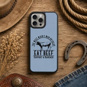 May include: A light blue phone case with a black border featuring the text "The West Wasn't Won On Salad" and "Eat Beef Support A Rancher" with a cow illustration. The case is on a wooden surface with a cowboy hat and horseshoe.