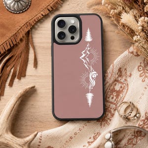 May include: A mauve phone case with a black border features a white graphic design of a mountain, sun, and tree. The case is surrounded by a brown leather bag with fringe, dried flowers, and jewelry on a wooden surface.