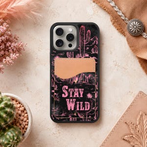May include: Black phone case with a pink desert landscape design featuring cacti and the words "STAY WILD." The case has a light brown cardholder on the back.