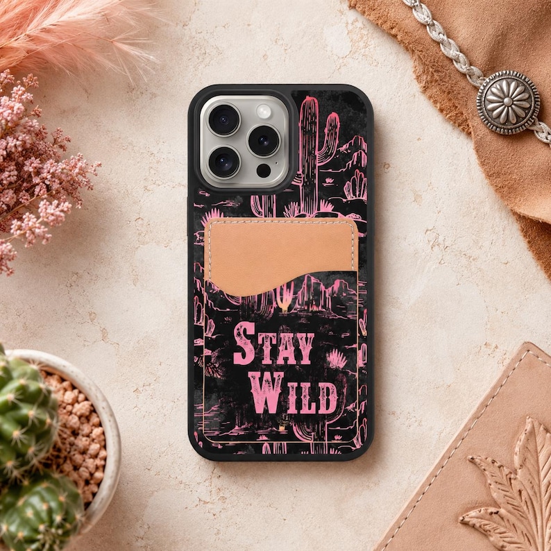 May include: Black phone case with a pink desert landscape design featuring cacti and the words "STAY WILD." The case has a light brown cardholder on the back.