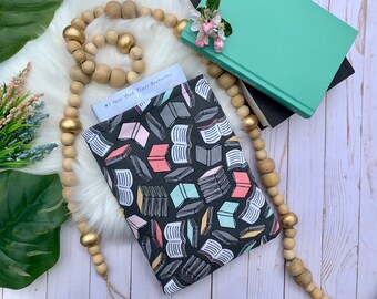 Puede incluir: Una funda de libro negra con un patrón de libros coloridos. La funda está hecha de tela y tiene una cremallera. La funda está sobre una superficie blanca con un libro y una cadena de cuentas de madera.