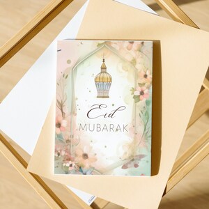 May include: A greeting card with a watercolor background featuring a floral design and a gold lantern. The text "Eid Mubarak" is written in brown calligraphy.