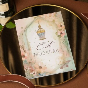 May include: A white card with a watercolor design of flowers and a gold lantern. The text "Eid Mubarak" is written in brown cursive script.
