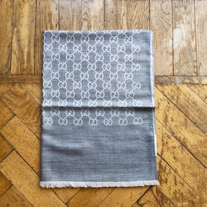 May include: A folded gray scarf with a white repeating pattern of interlocking "G" logos. The scarf has a fringed edge and is displayed on a wooden surface.