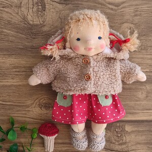 May include: A handmade doll with blonde yarn hair, blue eyes, and rosy cheeks. The doll wears a tan, button-up coat, a red polka-dot skirt with green pockets, and gray booties. A small, knitted mushroom is next to the doll.